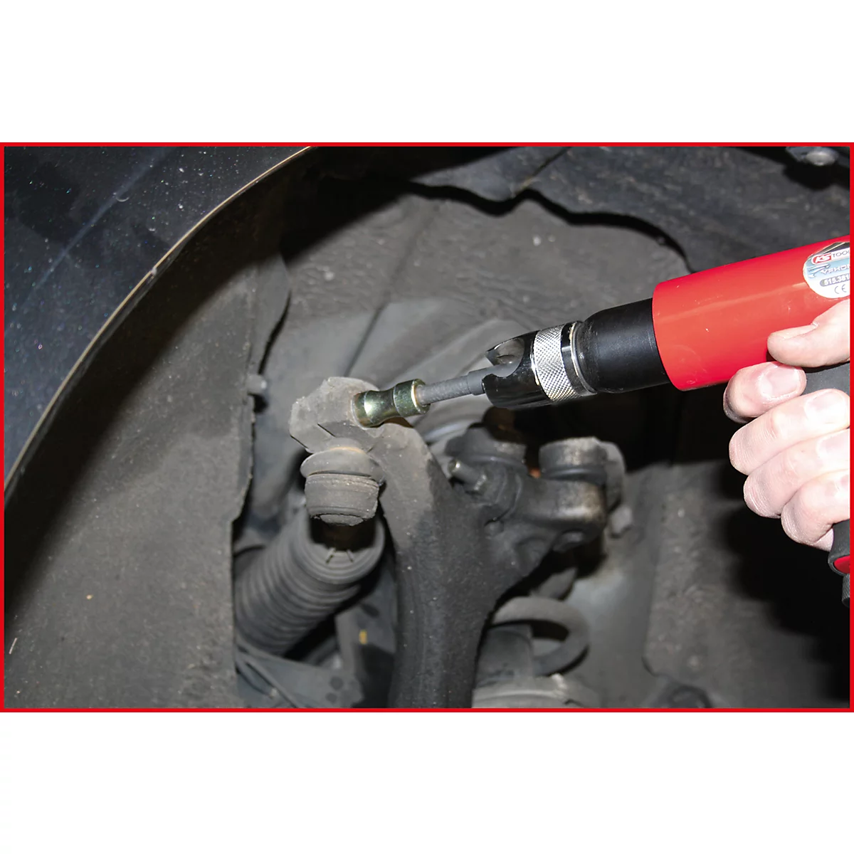 Nahaufnahme: Eine Hand bedient einen roten Druckluftschrauber an einem Fahrzeug-Stoßdämpfer.